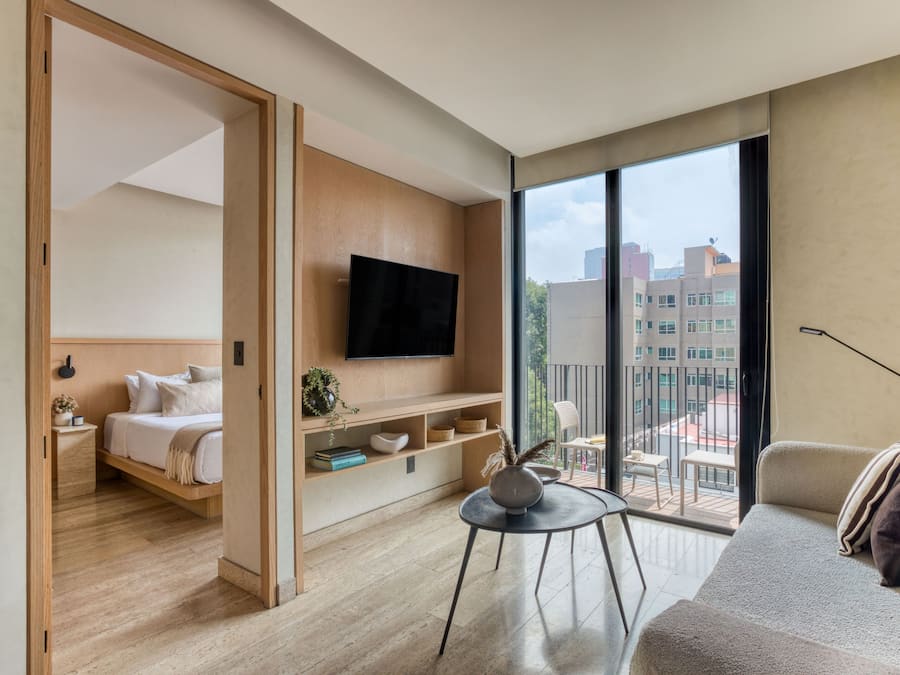 Una habitación de hotel moderna con una cama, un televisor montado en la pared, un balcón con vista a la ciudad y una zona de estar con mesa y sillas.