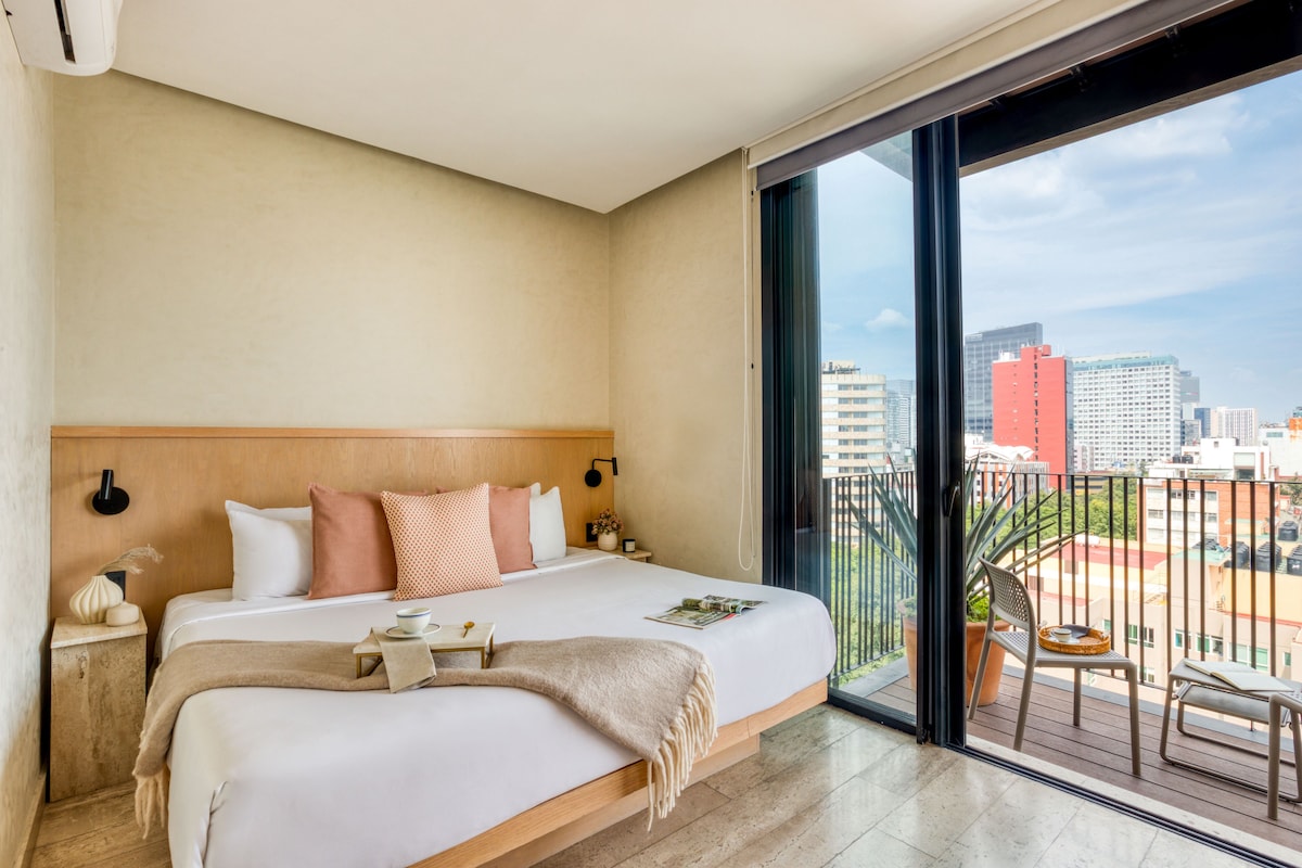 Quarto de hotel com uma cama grande, mesa de cabeceira, janela com vista para a cidade e varanda com cadeira e mesa.