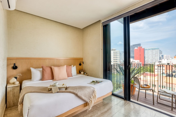 Quarto de hotel com uma cama grande, mesa de cabeceira, janela com vista para a cidade e varanda com cadeira e mesa.