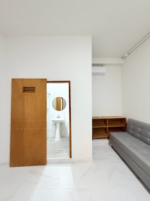 Een kamer met een houten deur, een wit wastafelmeubel, een spiegel, een grijze bank en een houten plank.