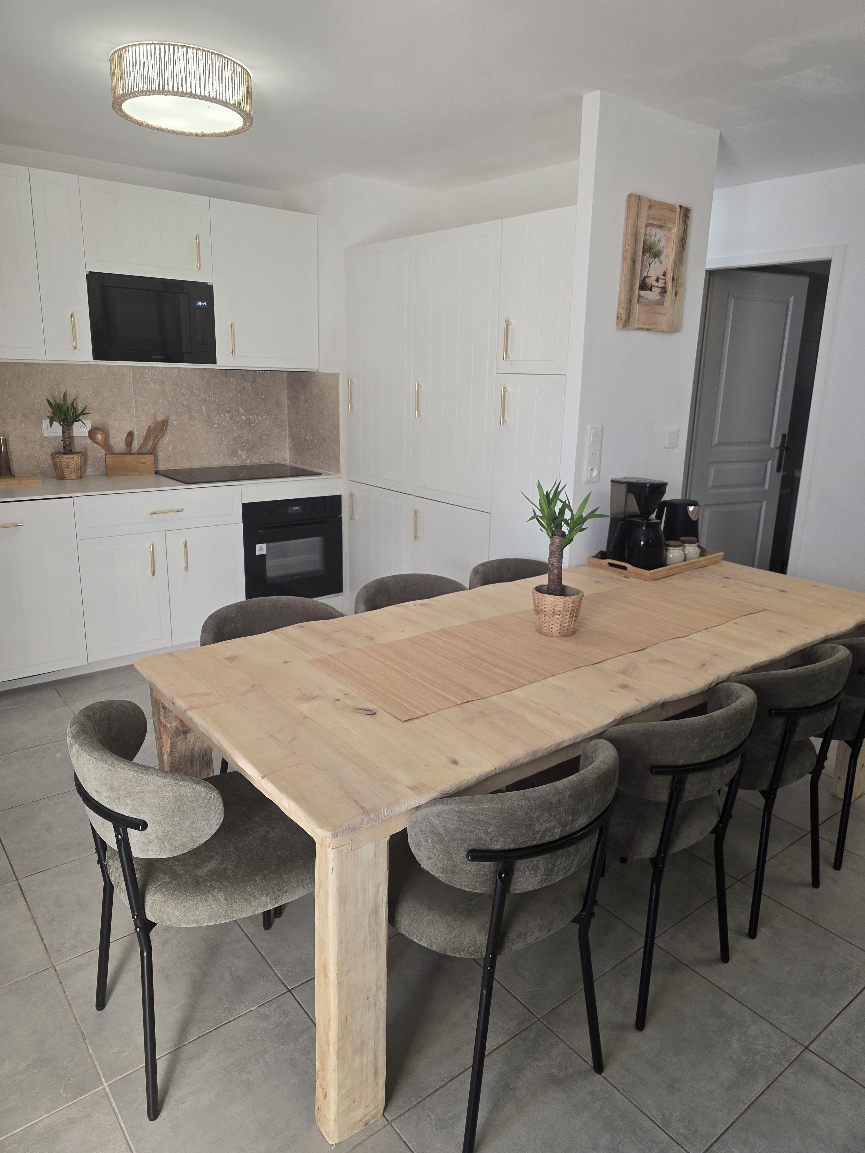 A dining area with a wooden table, grey upholstered chairs, a kitchen with white cabinets, and a ceiling light fixture.