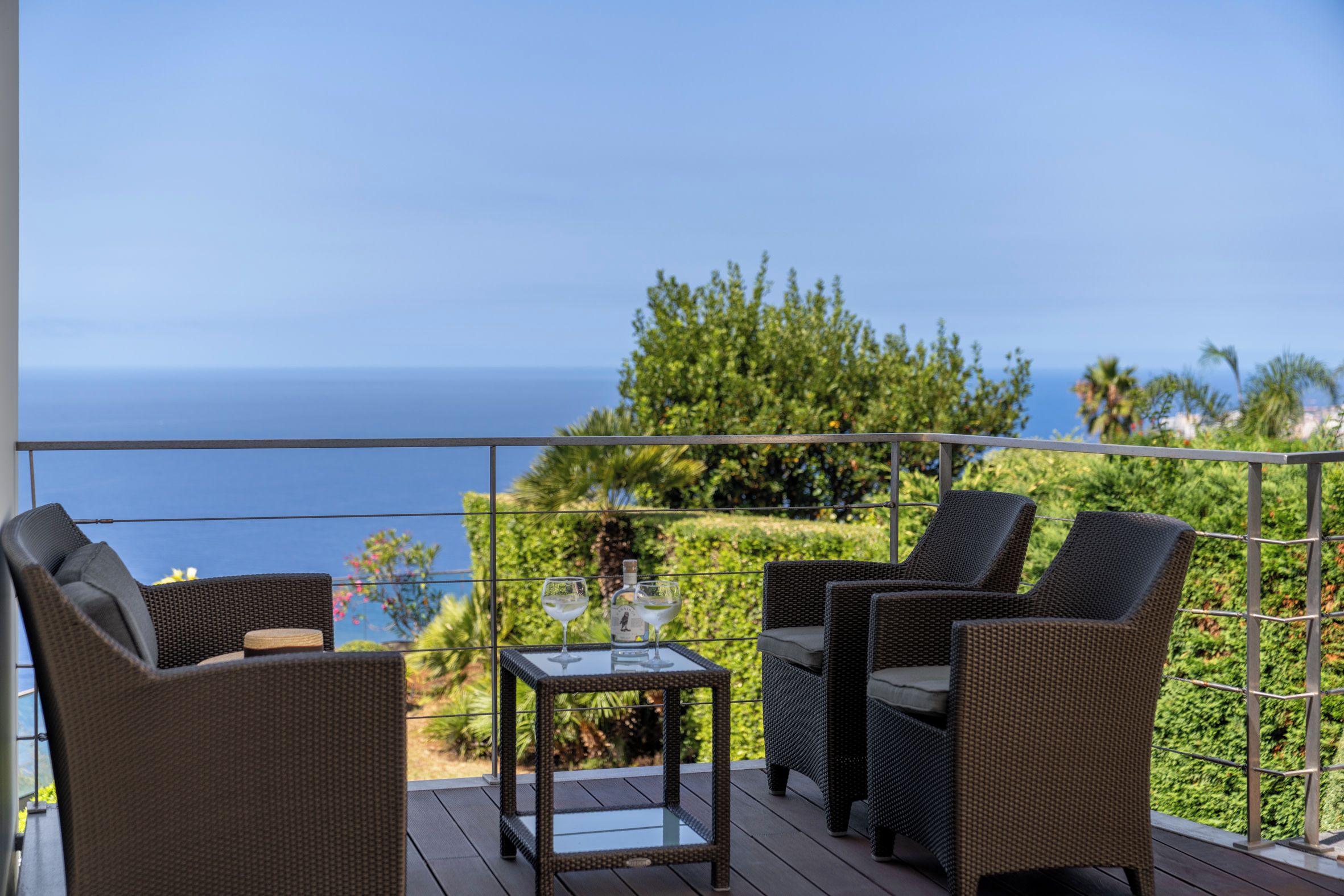 A balcony with wicker furniture and a glass table, overlooking a coastal view.