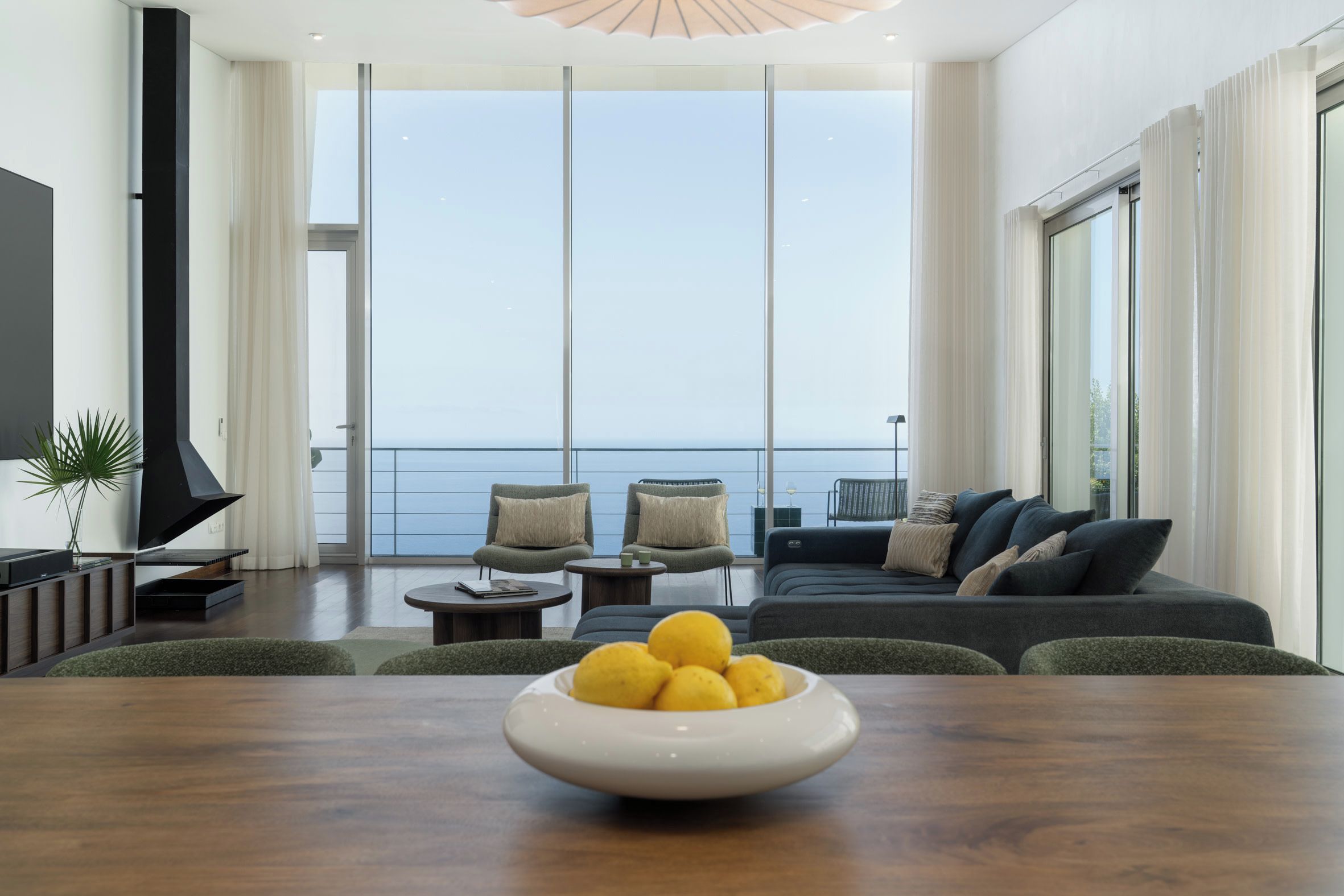 Ein modernes Wohnzimmer mit einem großen Fenster mit Meerblick, einer Sofagarnitur, einem Esstisch mit einer Schale Obst und einer kleinen Pflanze.