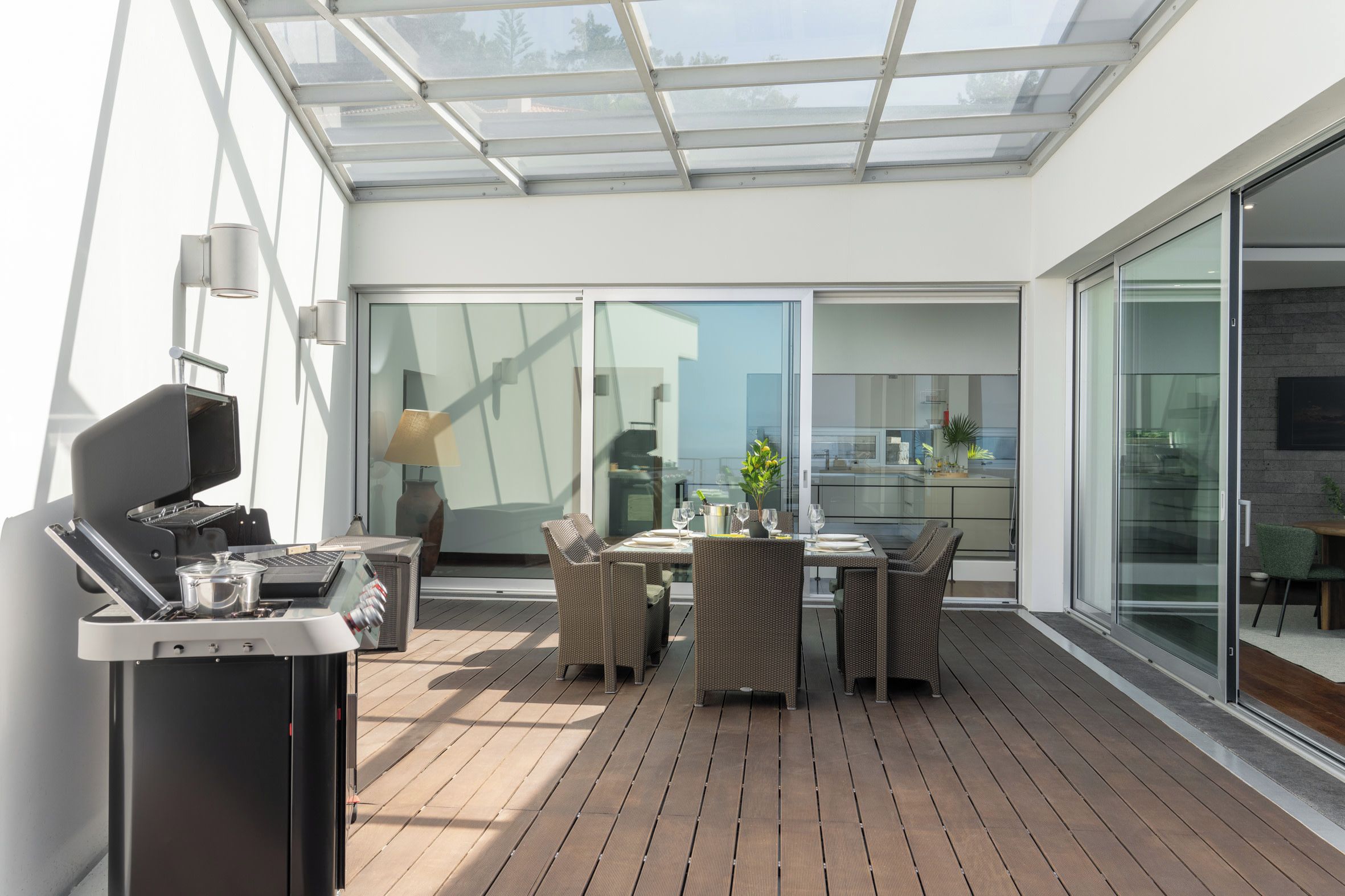 An outdoor patio with a grill, dining table, and chairs, a glass-enclosed area, and a view of the interior.