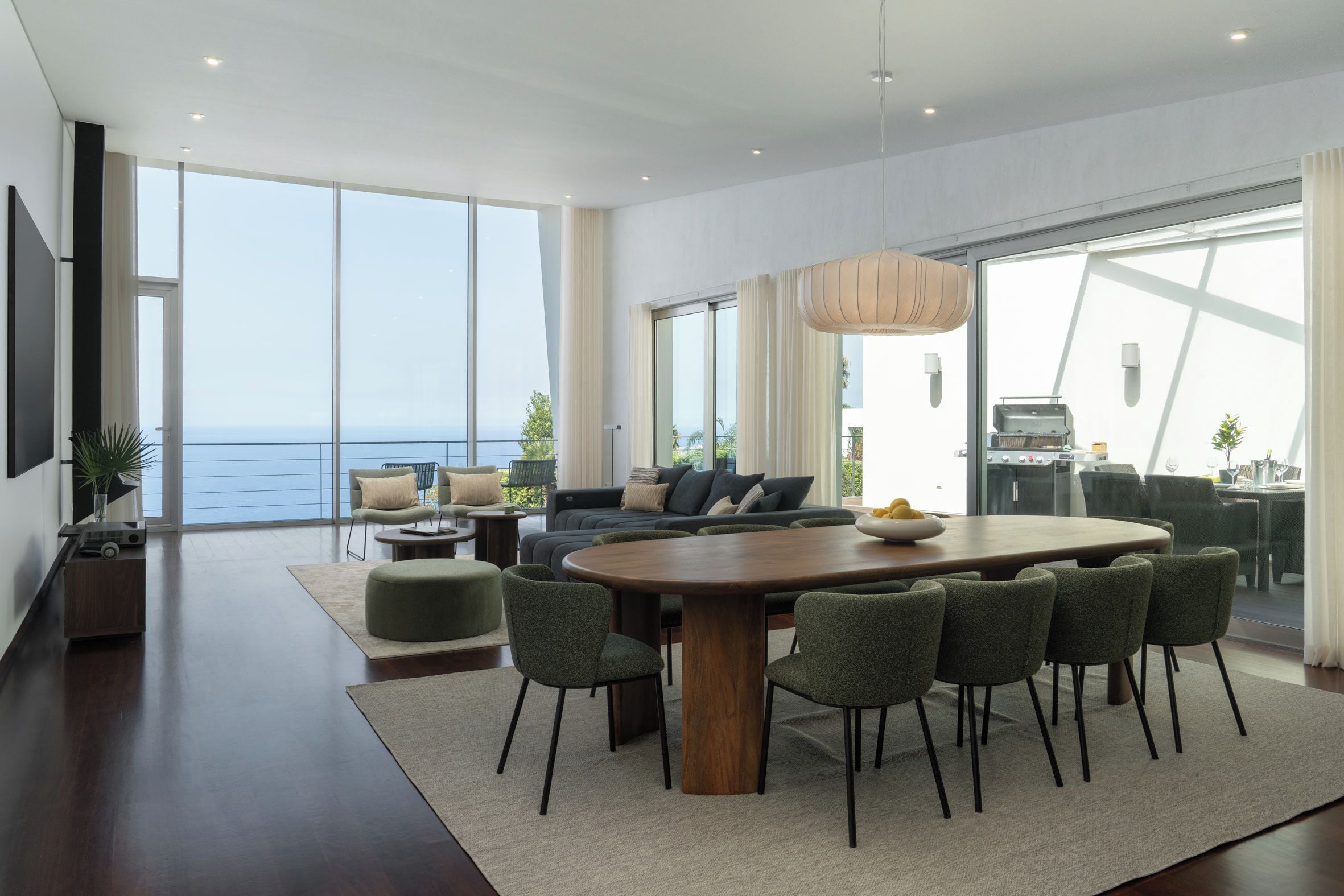 A modern living room with a large wooden dining table surrounded by chairs, a sofa, and a TV mounted on the wall.