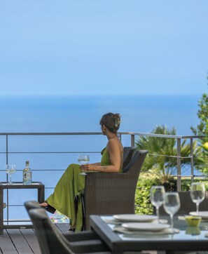Une personne est assise sur une chaise en osier, face à une étendue d’eau, avec une table et d’autres chaises au premier plan.