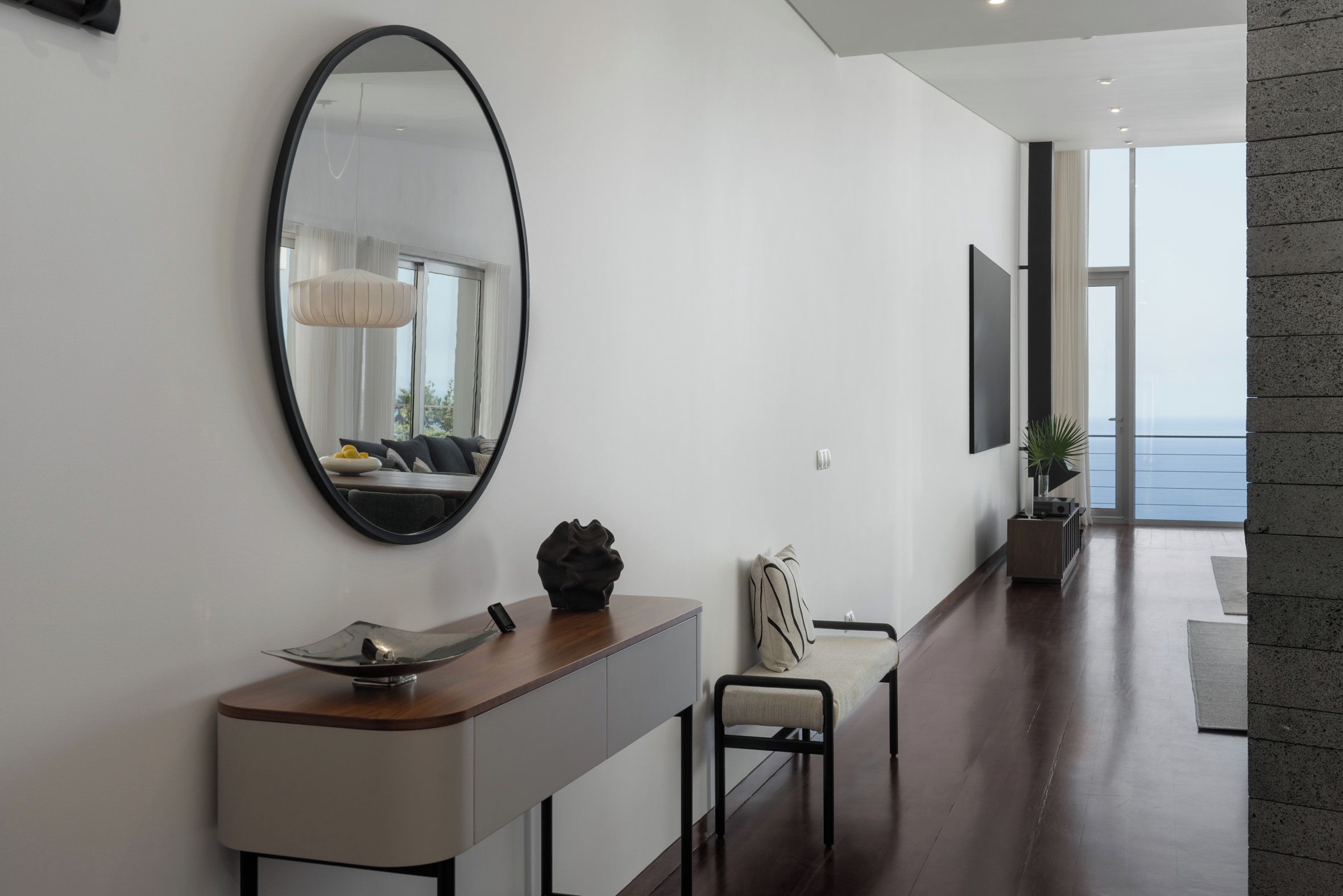Um interior moderno com um espelho oval grande, um console de madeira, uma cadeira e vista para o mar através de uma porta de vidro.