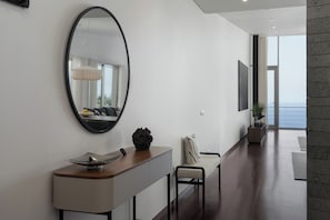 A modern interior with a large oval mirror, a wooden console table, a chair, and a view of the sea through a glass door.