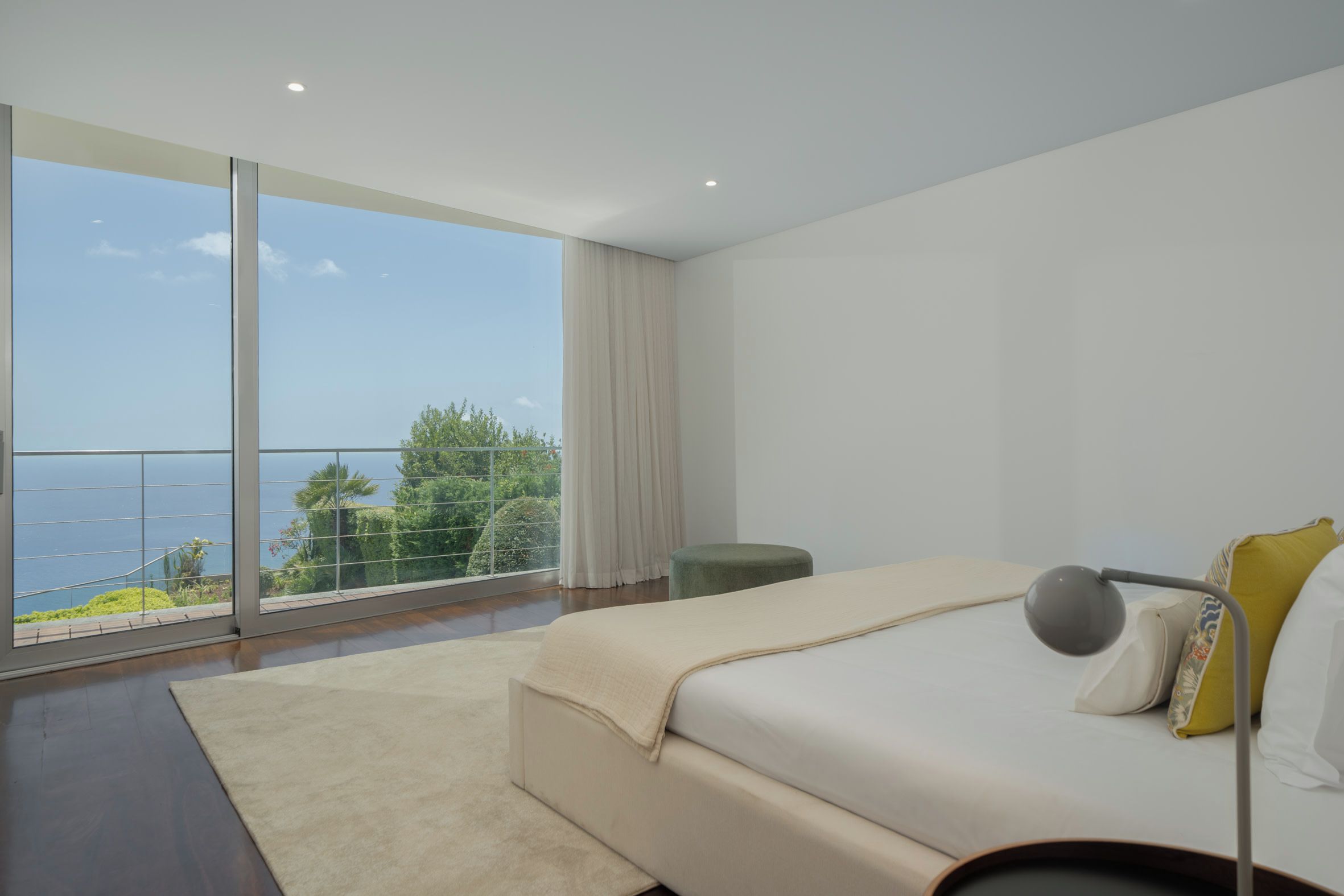 A modern bedroom with a large window, a bed, a bedside table, and a view of the sea.