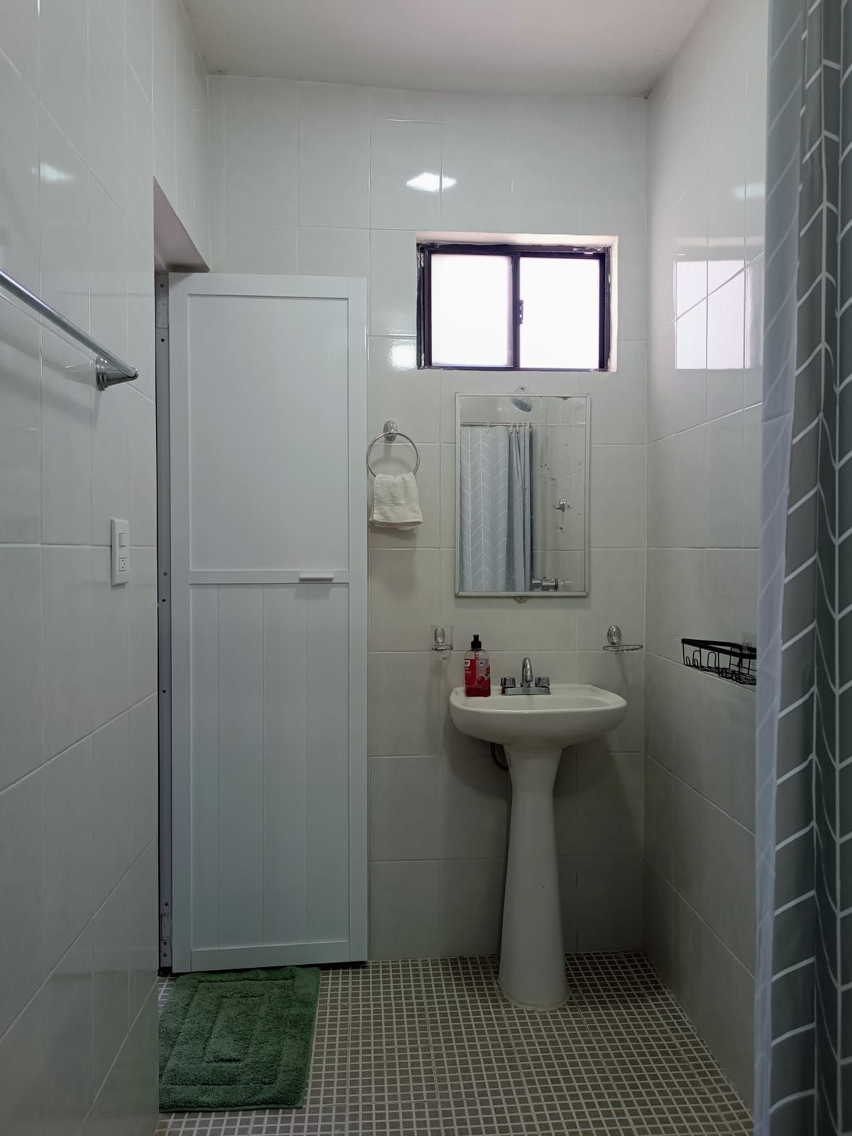 Un baño pequeño con una puerta blanca, una ventana, un lavamanos y un espejo.