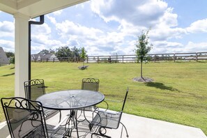 Eine überdachte Terrasse mit einem runden Metalltisch und Stühlen, einem kleinen Baum und einem umzäunten Garten.