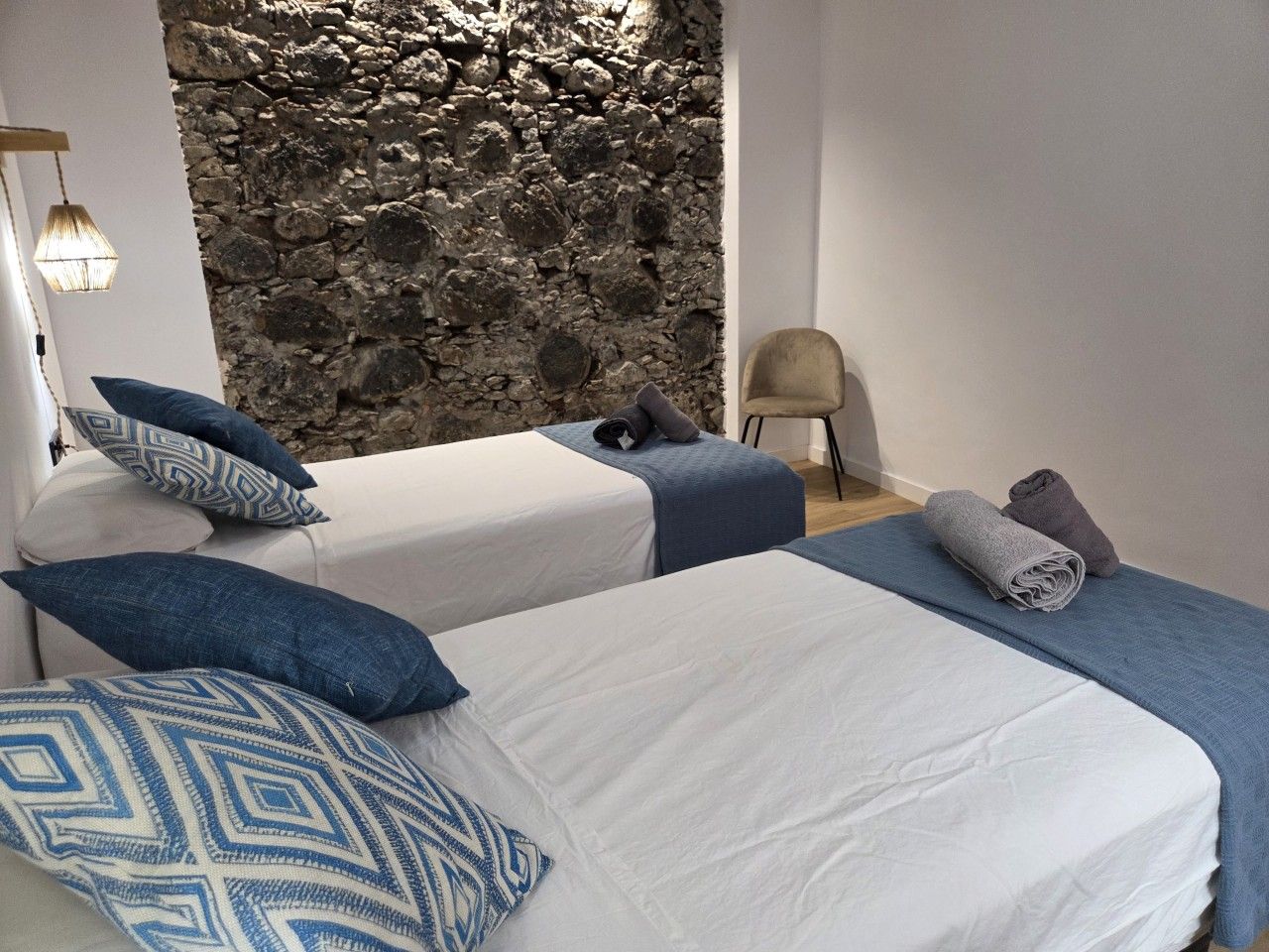 A bedroom with two beds, a stone wall, a chair, and a lamp.