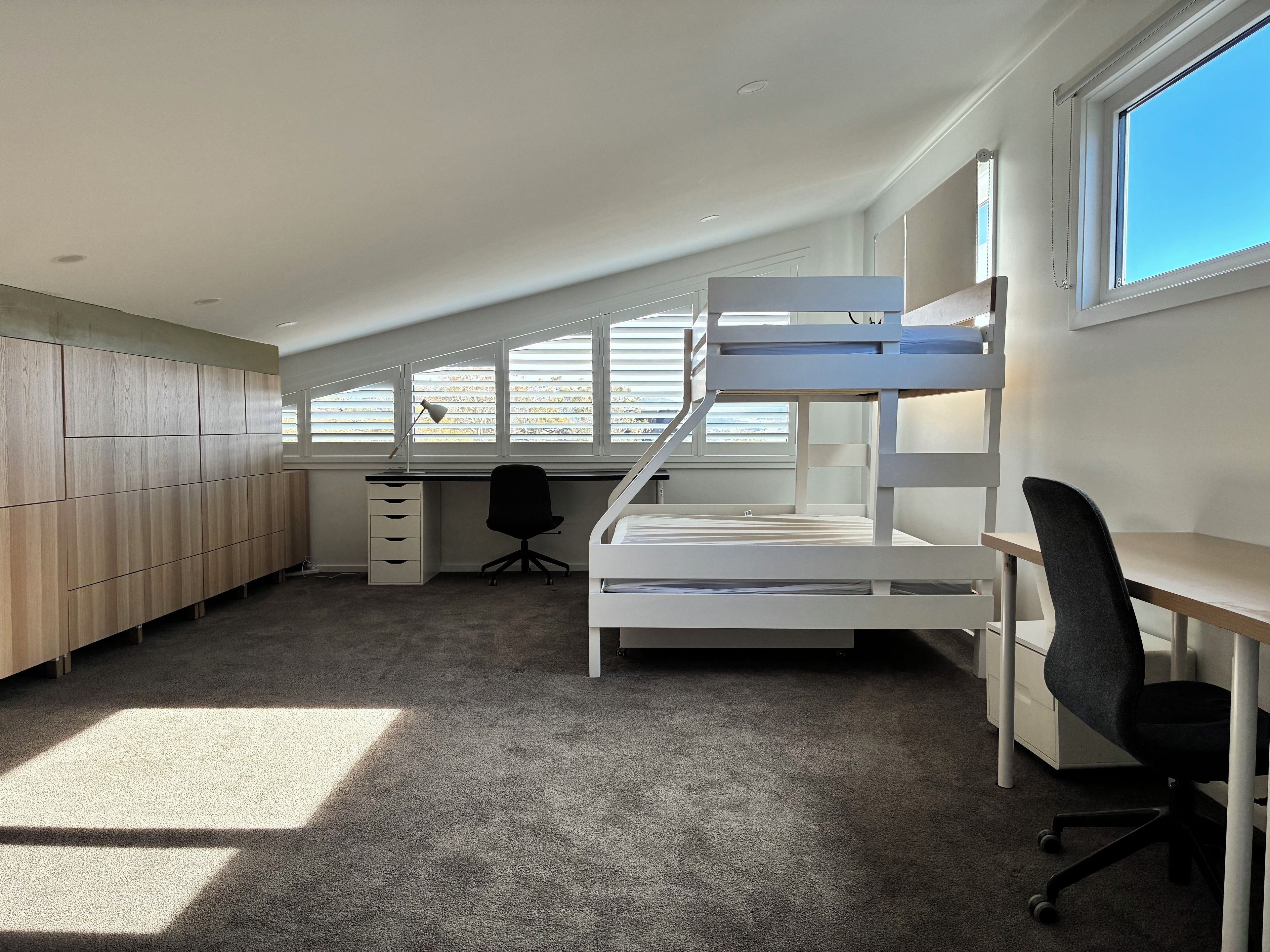 A bunk bed with a desk and chair, a wardrobe, and a window with blinds.