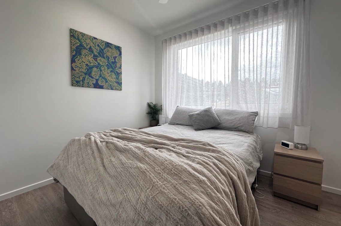 A bedroom with a bed, a nightstand, a lamp, a painting on the wall, and a window with curtains.