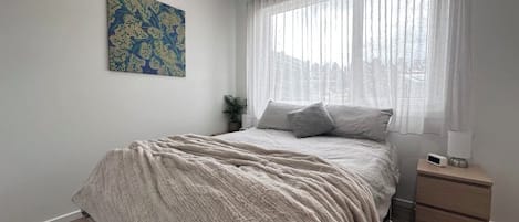 Une chambre à coucher avec un lit, une table de chevet, une lampe, un tableau au mur et une fenêtre avec des rideaux.