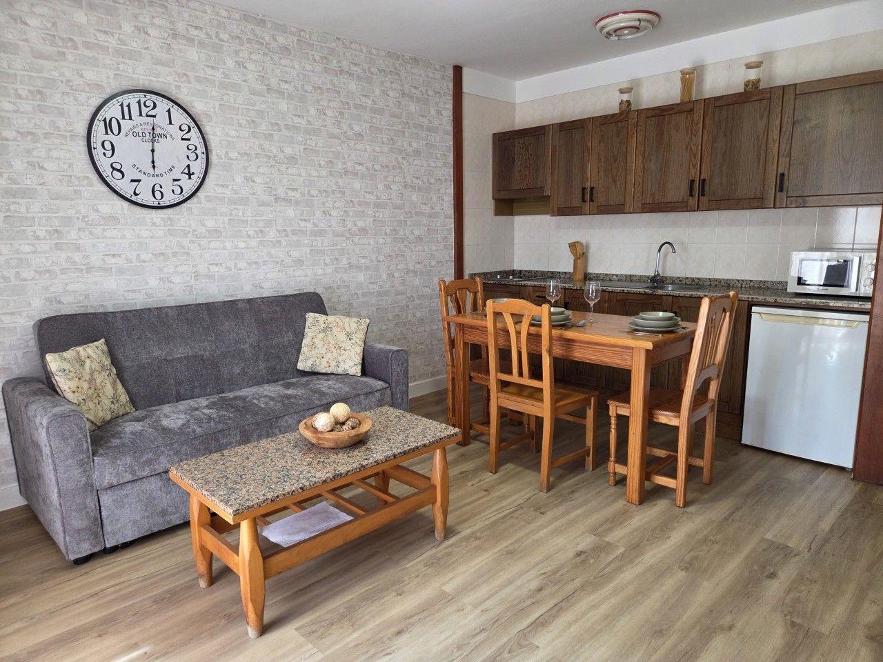 Un salon comprenant un canapé, une table basse et une table à manger avec des chaises. Il y a également un coin cuisine avec des placards en bois et un four à micro-ondes. Une horloge est fixée au mur.