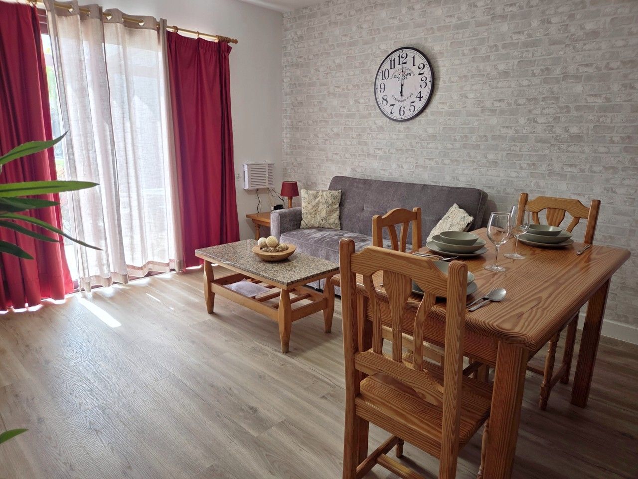 Un comedor con una mesa de madera dispuesta para cuatro personas, un sofá gris y un reloj en la pared.