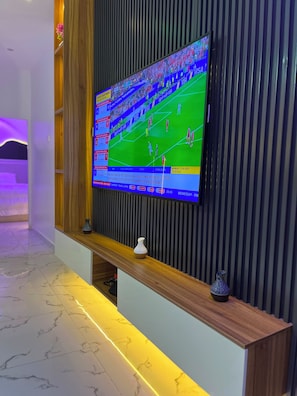 A flat-screen TV mounted on a wall with a wooden shelf below displaying two vases. - Flexxy Apartment Hotel (Osogbo)