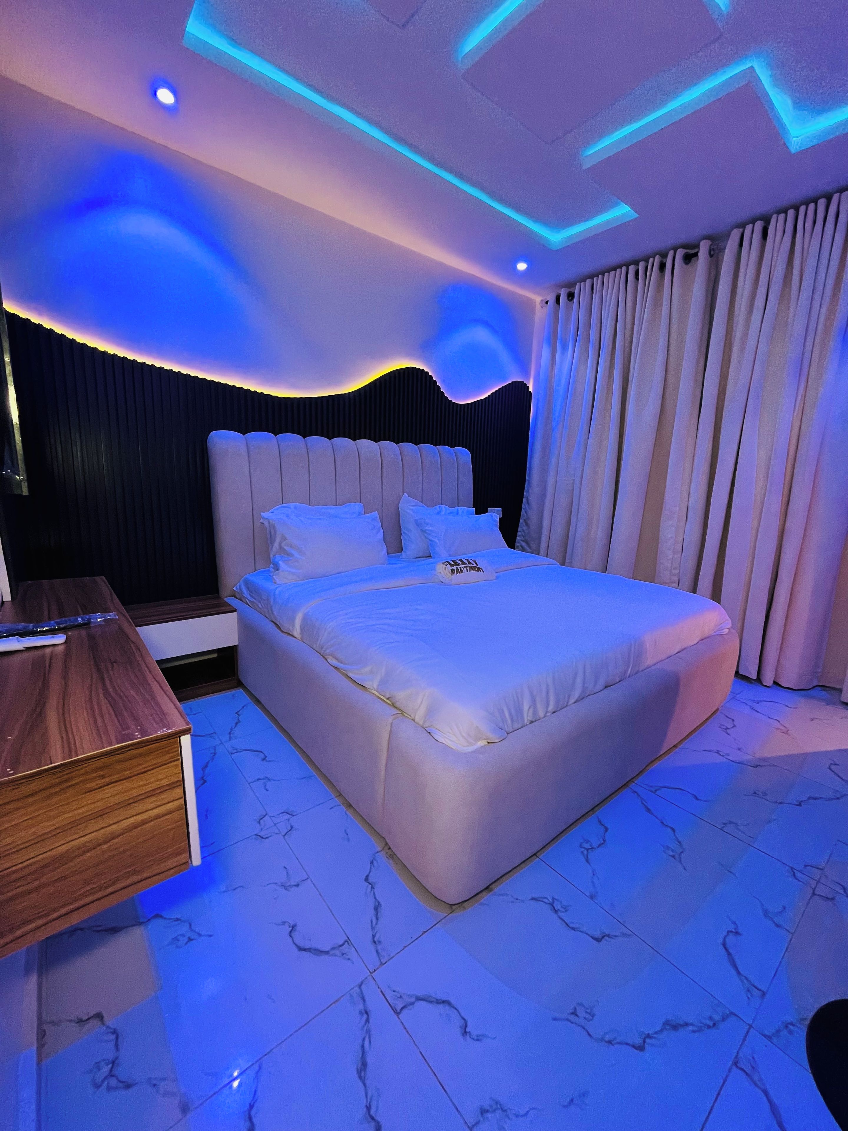 A modern bedroom with a large bed, a wooden nightstand, and a ceiling with blue lighting.