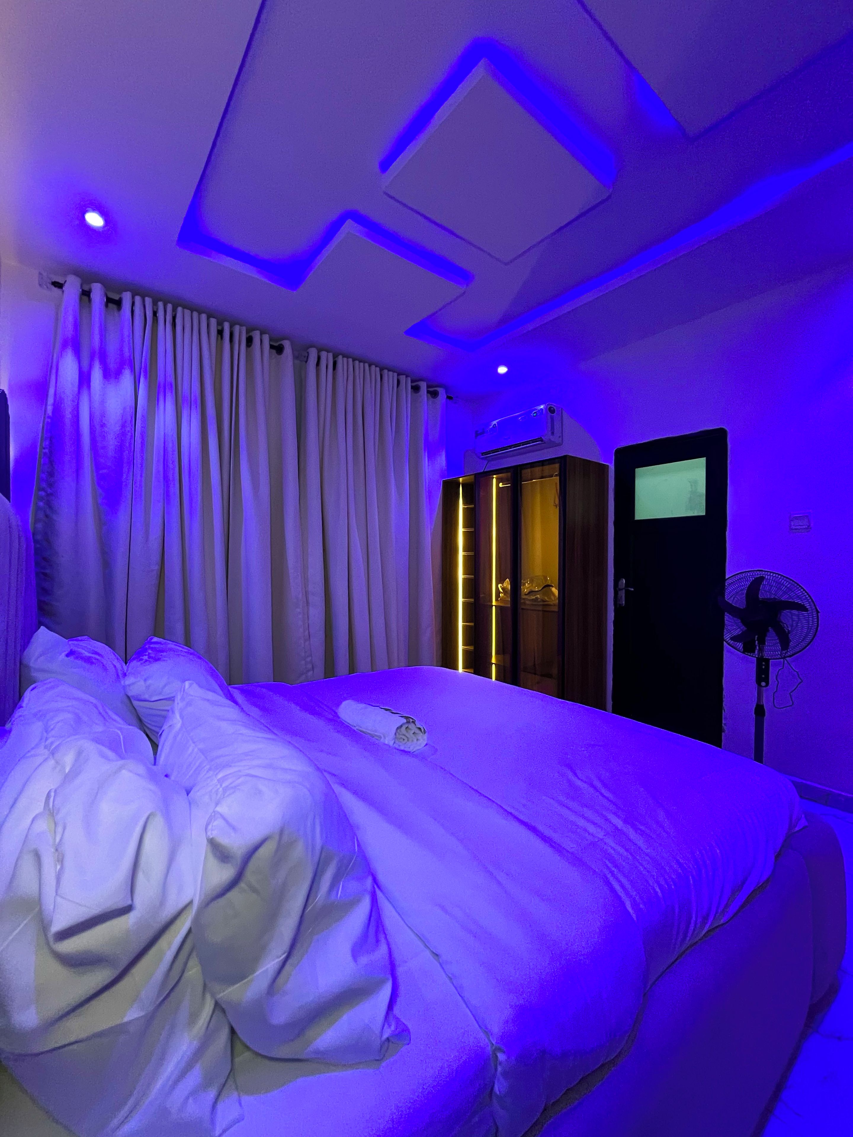 A hotel room with a bed, curtains, an air conditioning unit, a television, and a standing fan.