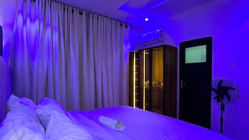 A hotel room with a bed, curtains, an air conditioning unit, a television, and a standing fan.