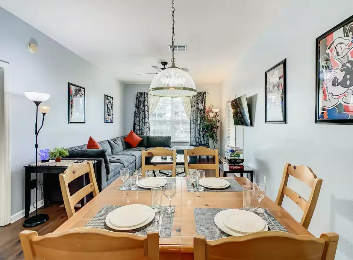 A dining area with a wooden table set for four, surrounded by chairs, with a ceiling fan above and framed artwork on the walls.