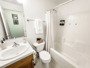Un baño con lavamanos, inodoro y bañera blancos. Hay un perchero con una toalla blanca y una cesta de artículos de aseo.