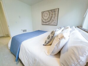 Un dormitorio con cama, una colcha azul, almohadas estampadas, una obra de arte en la pared y una ventana.