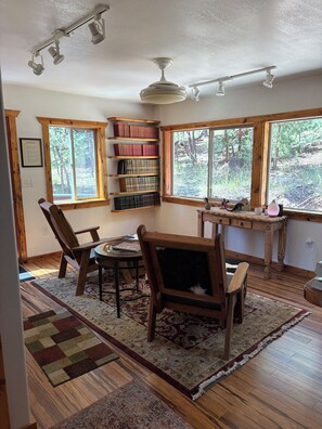 Ein Zimmer mit einem Holztisch, Stühlen, einem Bücherregal und großen Fenstern.