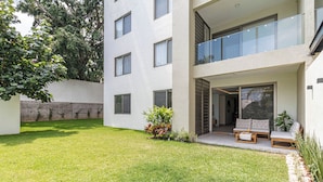 Een modern gebouw met twee verdiepingen, een plat dak, een balkon met glazen hekwerk en een kleine tuin met gras en potplanten.