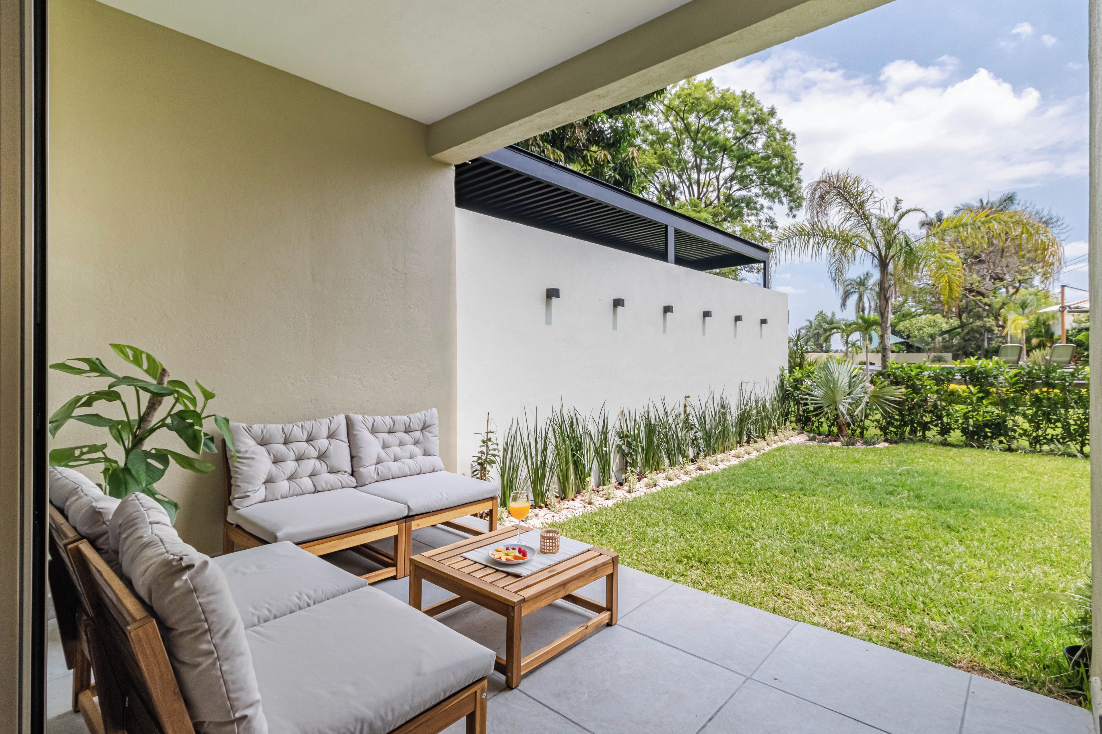 Un espacio al aire libre moderno con una zona de estar, un sofá y una mesa, una pared blanca con ventanas y un césped bien cuidado.