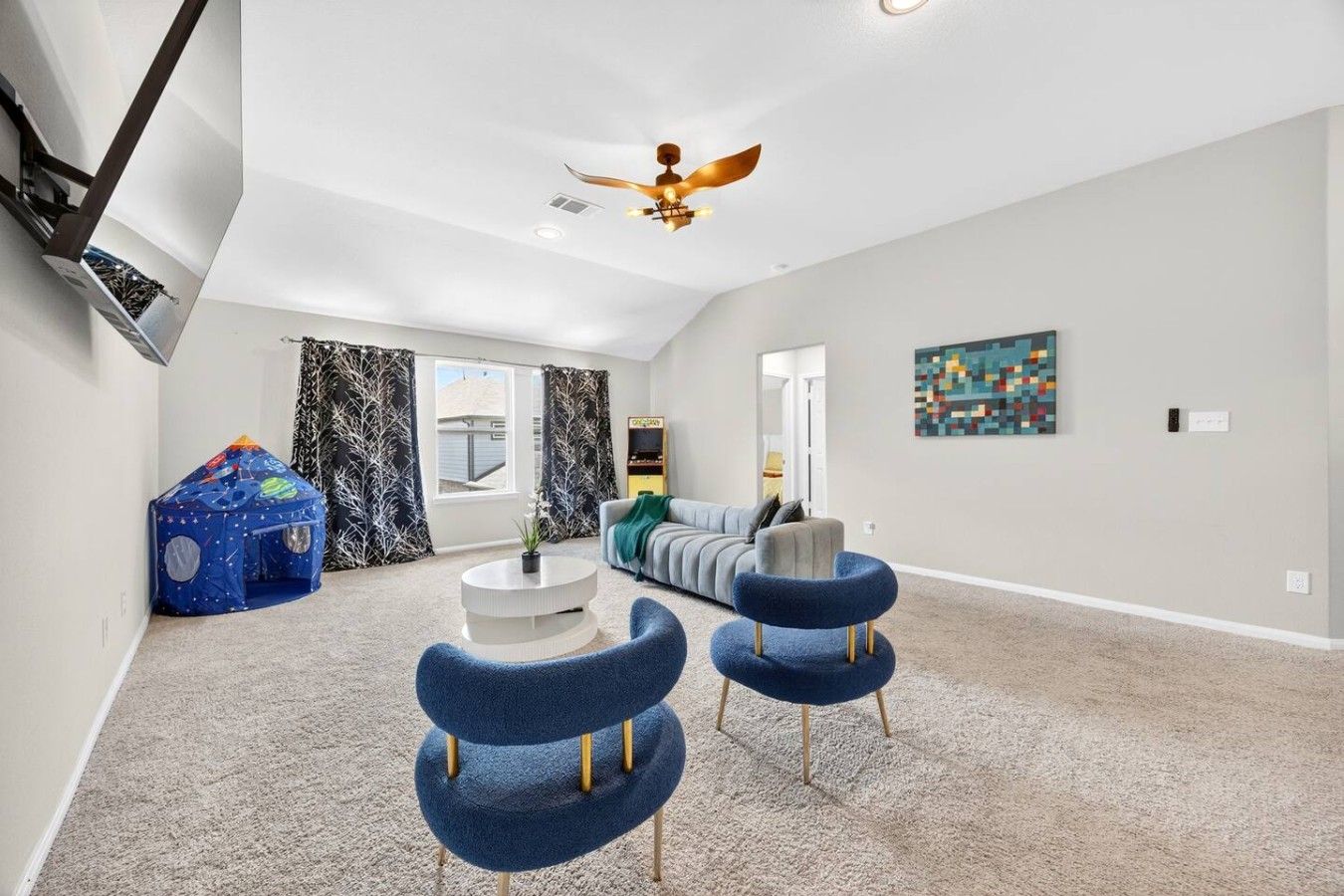 A spacious living room with a high ceiling, a ceiling fan, a sofa, two blue chairs, a small table, and a play tent.