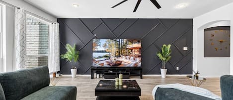 A modern living room with a large flat-screen TV, dark wall paneling, and a ceiling fan.