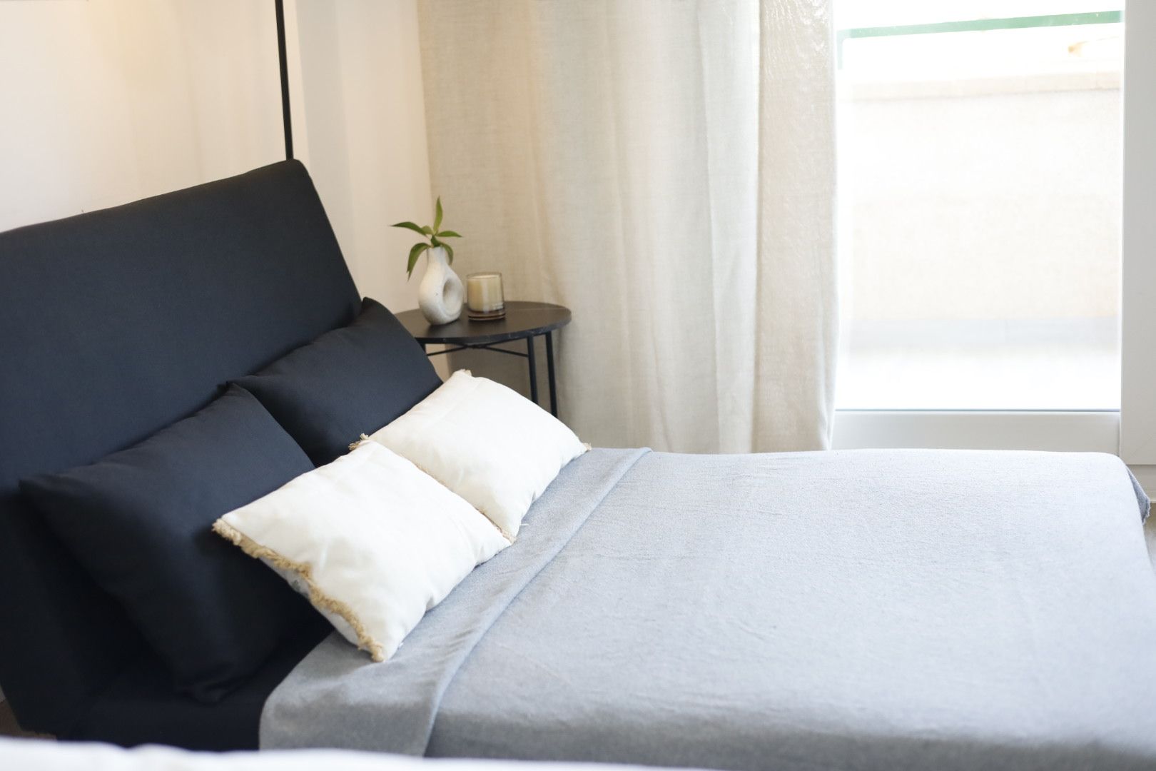 Un lit bien fait, avec une couvre-lit grise et deux oreillers, une petite table de chevet ornée d’un vase et d’une bougie, et une fenêtre avec des rideaux.