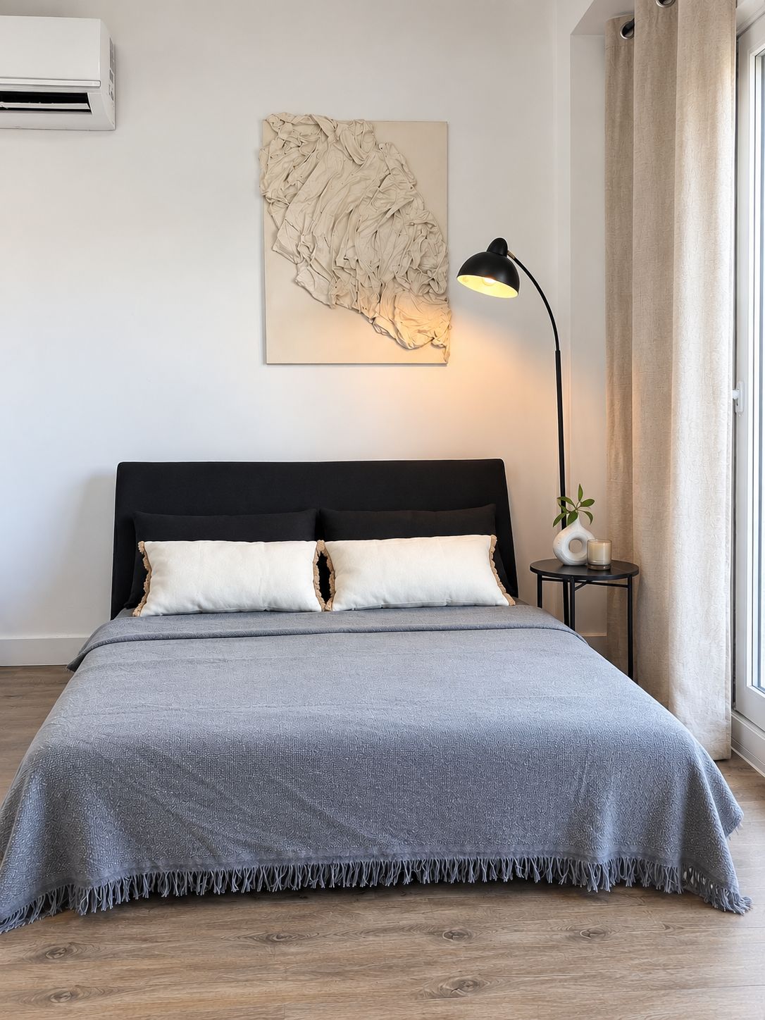 Un dormitorio con cama, aire acondicionado de pared, lámpara de pie, mesita con vela y ventana con cortinas.
