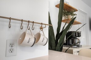 A kitchen with a metal rack holding white mugs, a potted plant, and a coffee maker on the counter. - Sunny Pinecraft Retreat (Sarasota)