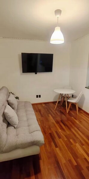 Ein Wohnzimmer mit einer Couch, einem an der Wand montierten Fernseher, einem runden Tisch mit Stühlen und einer Hängelampe.
