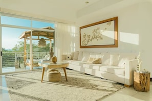 A spacious living room with a white sofa, a round coffee table, and a large window offering a view of greenery. - Ideally situated in the heart of town and a 4 min walk to the Beach. (Tamarindo)