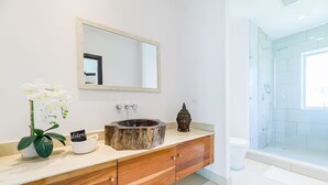 Un baño con tocador de madera, un lavamanos redondo, un espejo, zona de ducha y una planta pequeña.