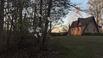 Une maison avec un toit en pente, entourée d’une cour verdoyante et d’arbres.