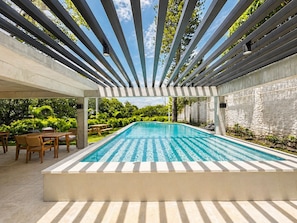 A rectangular pool with a clear blue water surface, surrounded by a pergola structure, and a seating area with tables and chairs. - Vrbo Property (San Salvador)