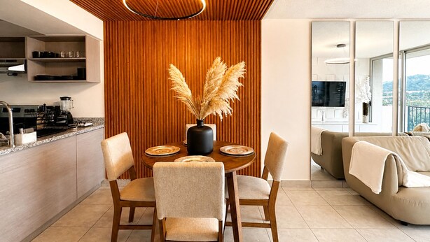 Une cuisine moderne avec un lambris en bois, un coin repas avec une table ronde et des chaises, et un salon avec un canapé et une vue sur l’extérieur.