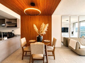 A modern kitchen with wooden paneling, a dining area with a round table and chairs, and a living room with a sofa and a view of the outdoors.