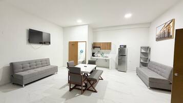 Una sala de estar moderna con un sofá gris, una mesa de comedor con sillas y una cocina con nevera y armarios.