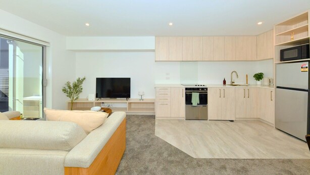 Un apartamento moderno con sala de estar, televisor de pantalla plana, sofá, cocina con armarios de madera clara, horno empotrado y refrigerador.