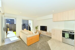 A modern living room with a sofa, TV, and kitchen area with cabinets and a stove. - Stylish 1 Bedroom Apartment with Private Yard (Christchurch)
