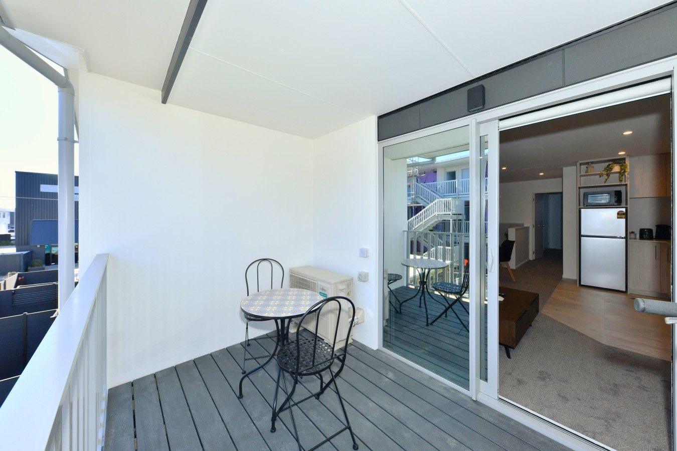 A balcony with a table and chairs, a sliding glass door leading to a staircase, and a kitchen area with a refrigerator.