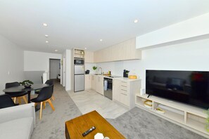 Uma sala de estar moderna com televisão de tela plana, uma área de jantar com mesa e cadeiras, e uma cozinha com armários e eletrodomésticos em tons claros.