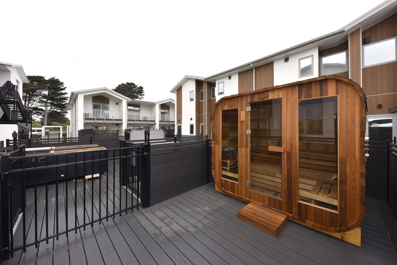 Uma sauna de madeira num deck com uma cerca de metal preta, rodeada por edifícios modernos.