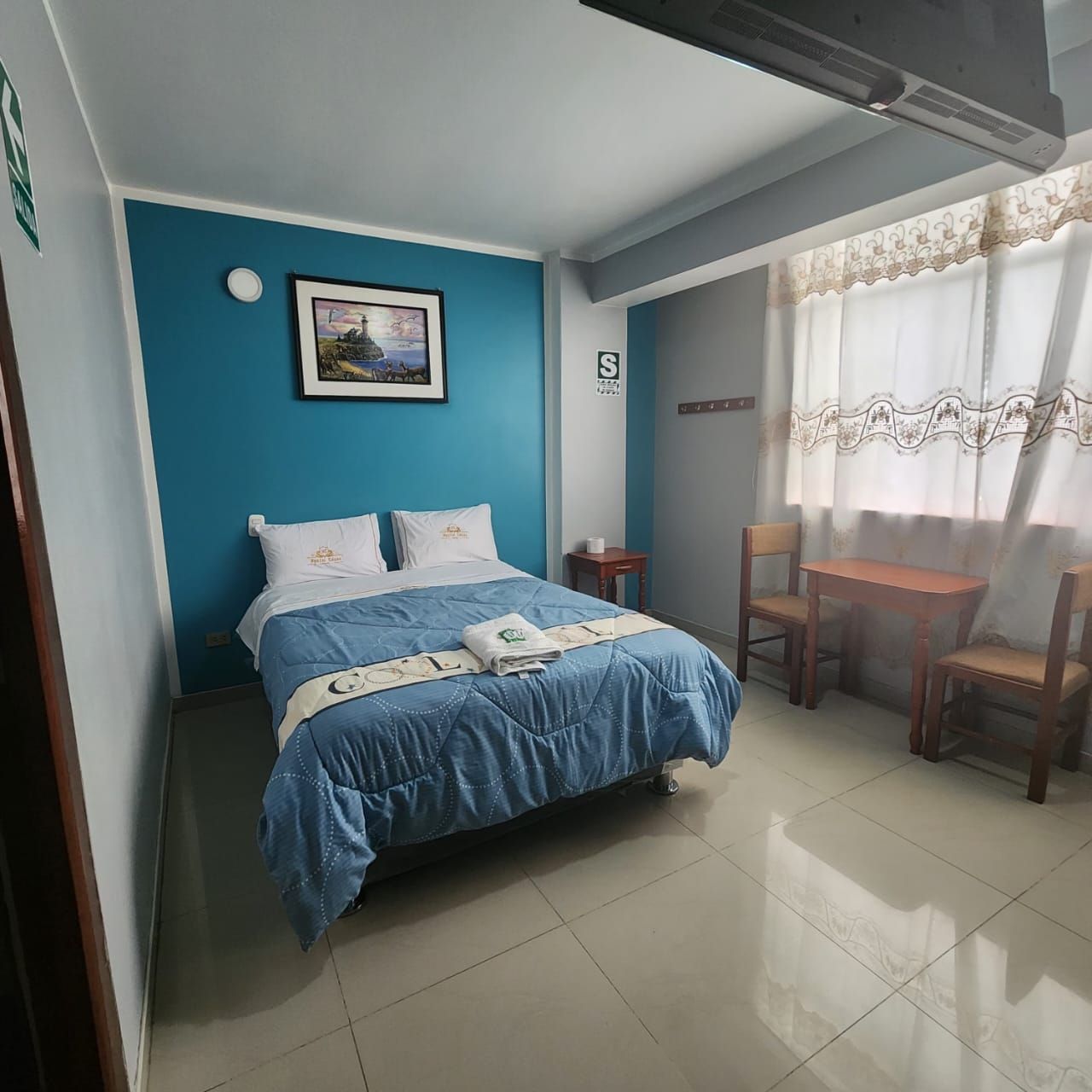 A bedroom with a bed, a small table, two chairs, a picture on the wall, and a window with curtains.